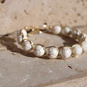 Puede incluir: Una pulsera dorada con cuentas blancas estampadas. Las cuentas están unidas con un fino alambre dorado, creando un accesorio elegante. Esta pulsera es ideal para añadir un toque de sofisticación a cualquier atuendo. Longitud: aproximadamente 18 cm.