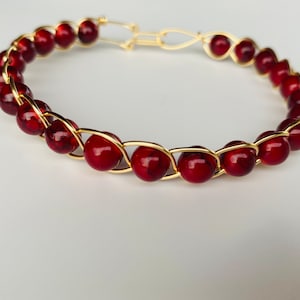May include: A gold wire wrapped bracelet with red beads. The bracelet is made with a simple wire wrapping technique and features a clasp closure.