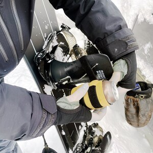 May include: A black snowboard with a black and yellow can holder. The can holder has the text "ROME SDS ROME" on it. The snowboard is on a snowy surface.