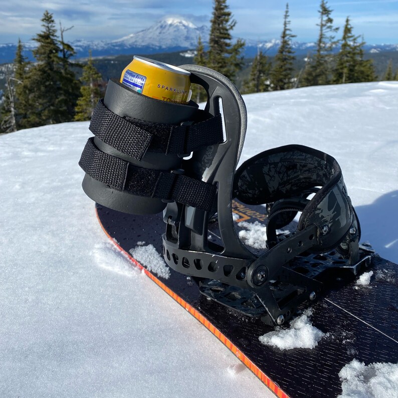 May include: A black snowboard binding with a built-in can holder. The holder is made of black fabric and has a velcro strap to secure a can. The binding is mounted on a black snowboard.