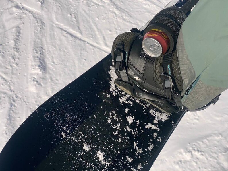 May include: A black snowboard with a black binding and a can of soda attached to the binding.