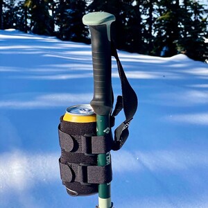 May include: A black and green hiking pole with a black and grey can holder attached to it. The can holder has adjustable straps and is holding a yellow can.