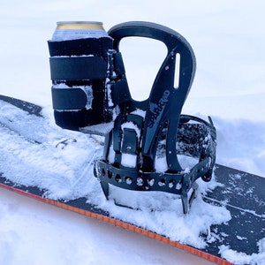 May include: A black snowboard binding with a can holder attached to the side. The can holder is made of black fabric and has a strap to secure a can of beer. The binding is sitting on a snowboard in the snow.