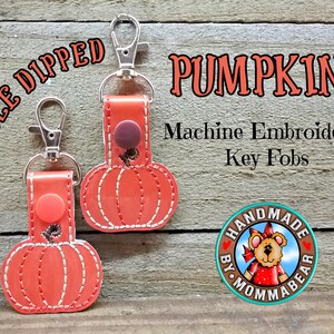 May include: Two orange pumpkin-shaped key fobs with white stitching and silver hardware. The text "PUMPKINS" and "Machine Embroidered Key Fobs" are visible. The image also includes the text "DOUBLE DIPPED" and a "HANDMADE BY MOMMABEAR" logo.