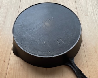 Puede incluir: Una sartén de hierro fundido negro con un mango largo y una superficie de cocción circular. La palabra "WAPAK" está grabada en la parte inferior, junto con el número "11". La sartén está sobre una superficie de madera clara.