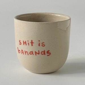 Puede incluir: Una taza de cerámica beige con bordes redondeados. Las palabras "shit is bananas" están escritas a mano en rojo en la parte delantera de la taza. La taza está vacía y se encuentra sobre un fondo blanco liso.