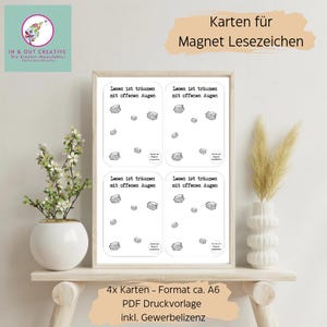 May include: A framed print with four cards, each displaying the text "Lesen ist träumen mit offenen Augen" and book illustrations. The print also includes the text "4x Karten - Format ca. A6 PDF Druckvorlage inkl. Gewerbelizenz". The image is on a wooden shelf with a white vase and plant.