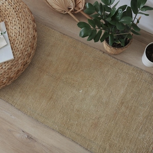 May include: A rectangular, beige woven rug with a textured surface. The rug is placed on a light-colored wooden floor, alongside a woven circular ottoman, potted plants, and a small cactus in a white pot. The rug's neutral color complements the natural elements.
