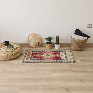 Op de afbeelding: Een rechthoekig tapijt met een centraal rood en beige geometrisch ontwerp, omlijst door een donkerblauwe rand en beige randen. Het tapijt ligt op een lichte houten vloer, met andere decoratieve items op de achtergrond.