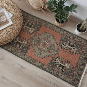 May include: A rectangular area rug featuring a terracotta orange background with a central geometric medallion in sage green and cream. The rug displays intricate symmetrical patterns, dark green borders, and short white fringe on both ends.