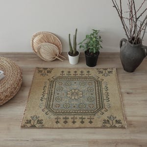 May include: A square area rug featuring a central floral medallion in light blue and beige, surrounded by a geometric border with repeating flower motifs in muted tones of brown, gray, and cream, finished with a short fringe on the edges.