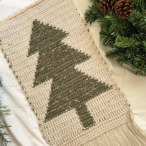 Puede incluir: Un tapiz de pared tejido a crochet con un diseño de árbol de Navidad verde sobre un fondo blanco. El tapiz tiene una varilla de madera en la parte superior y una franja en la parte inferior.