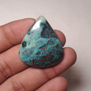 May include: A polished, teardrop-shaped gemstone. The stone displays a mix of teal, brown, and blue hues, creating a marbled effect. The gemstone is held in a hand, highlighting its size and form. This is a natural stone.