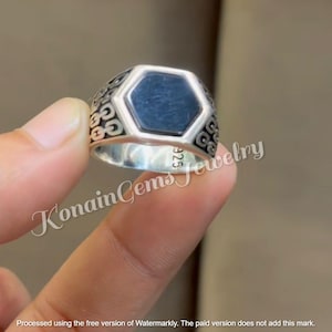 Può includere: Un anello d'argento con una pietra esagonale blu scuro. L'anello ha un design ornato sui lati e il testo "925" è visibile. L'anello è tenuto dalle dita di una persona. Il testo "Konain Gems Jewelry" è visibile.