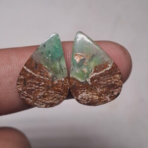 Puede incluir: Dos piedras preciosas en forma de lágrima. La parte superior de cada piedra es de un verde translúcido, que pasa a una base marrón y texturizada. Las piedras tienen una superficie pulida y reflectante, que muestra patrones y variaciones de color naturales.