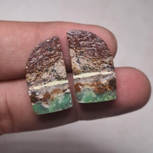 Puede incluir: Dos piedras preciosas semicirculares con apariencia en capas. La sección superior es marrón con un patrón texturizado, seguida de una banda color crema, una capa marrón y una base verde. Estas piedras podrían usarse para hacer joyas.