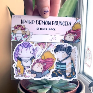 May include: A pack of eight stickers featuring cartoon cats in different outfits. The packaging reads "KPAWP DEMON POUNCERS STICKER PACK". The stickers are colourful and detailed, with various poses and hairstyles. The image is held in front of a window.
