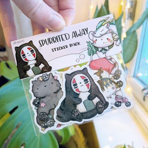 Może przedstawiać: Opakowanie ośmiu naklejek z postaciami z "Spirited Away". Naklejki przedstawiają czarną postać przypominającą kota z czerwoną maską, białego smoka i inne fantazyjne wzory. Opakowanie trzymane jest w dłoni.