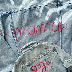 May include: Two denim jackets, one larger than the other, with the word "mama" embroidered in pink and red on the back of the larger jacket and the word "gigi" embroidered in pink and red on the back of the smaller jacket.