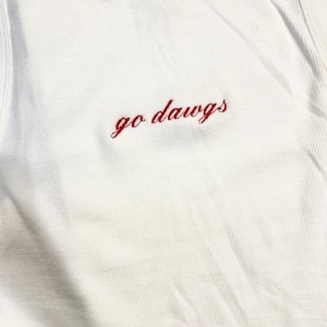 May include: White ribbed tank top with the embroidered phrase "go dawgs" in red script. The top has a classic, casual style, perfect for warm weather or layering.