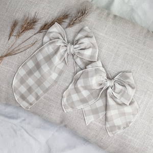 May include: Two beige and white gingham ribbon bows on a light beige fabric background.