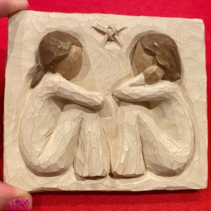 Könnte beinhalten: Eine weiße Keramik-Wandplakette mit einem geschnitzten Design von zwei Figuren, die Rücken an Rücken sitzen. Die Figuren haben braunes Haar und halten sich an den Händen. Ein kleiner Schmetterling ist über den Figuren geschnitzt.