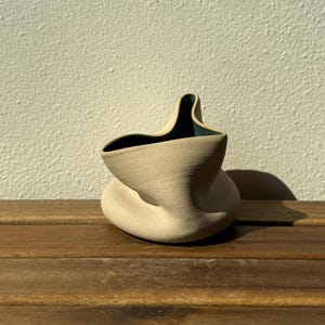 Op de afbeelding: Een kleine, sculpturale keramische vaas met een onregelmatige, gevouwen en organische vorm. De buitenkant heeft een matte, lichtbeige, getextureerde afwerking, terwijl de binnenkant een diepe, contrasterende donkergroenblauwe kleur heeft.