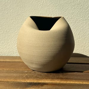 Könnte beinhalten: Eine kugelförmige Keramikvase mit matter beiger Außenseite und glatter, dunkler Innenseite. Das Gefäß zeichnet sich durch einen eingedrückten Rand aus, der eine tiefe Vertiefung an der Oberseite erzeugt und eine runde, organische Form bildet.