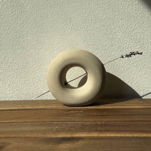 Könnte beinhalten: Eine kreisförmige, beige Keramikvase mit glatter, matter Oberfläche und einer zentralen Öffnung. Ein einzelner, schlanker Zweig getrockneter Lavendel ist horizontal positioniert und führt durch das mittlere Loch des ringförmigen Gefäßes.