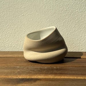 Könnte beinhalten: Ein kleines, asymmetrisches Keramikgefäß mit einer texturierten, matten beigen Außenseite und einer glatten, weiß glasierten Innenseite. Das Objekt hat eine markante, organisch geformte, eingedrückte Gestalt mit schrägem Rand und feinen Linien.