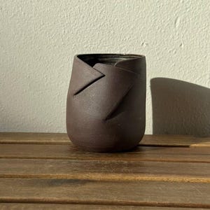 Könnte beinhalten: Ein dunkelbrauner Keramikbecher mit matter Oberfläche und einem gefalteten, überlappenden Randdesign. Das Gefäß hat einen abgerundeten Boden und eine glatte, strukturierte Oberfläche, die das Licht einfängt und subtile Schatten wirft.