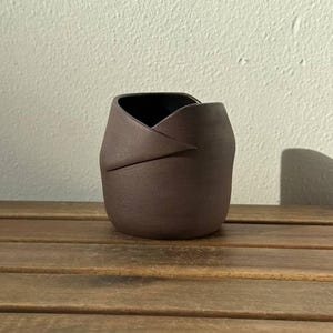 Könnte beinhalten: Ein dunkelbraunes Keramikgefäß mit matter Oberfläche und einem einzigartigen, gefalteten oder überlappenden Randdesign, das eine asymmetrische Silhouette erzeugt. Der Becher hat einen abgerundeten Boden und eine glatte, einfarbige Außenfläche.