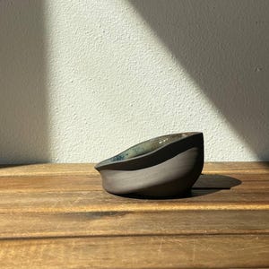 Könnte beinhalten: Eine kleine, handgeformte Keramikschale mit einer matten, dunkelgrauen Außenseite und einer glatten, glänzenden Innenseite mit gesprenkelter blau-grüner Glasur. Die Schale hat einen asymmetrisch geschwungenen Rand und einen abgerundeten Boden.