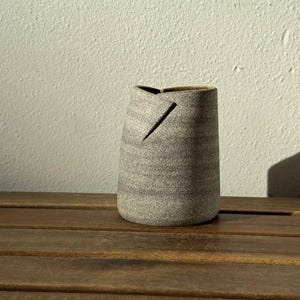 May include: A cylindrical ceramic vessel with a speckled light gray and beige finish. The top edge features a distinct diagonal slit detail, creating an asymmetrical opening. The surface has a textured appearance with subtle horizontal banding.