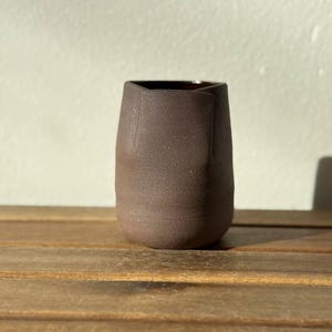 Könnte beinhalten: Ein dunkelbrauner Keramikbecher mit einer texturierten, matten Außenfläche. Das Gefäß zeichnet sich durch ein geometrisches Randdesign mit nach innen gefalteten Kanten aus, die eine markante Silhouette bilden. Das Innere ist glatt und dunkel, was einen Kontrast zur rauen Oberfläche bildet.