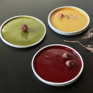 May include: Three small ceramic dishes with white rims. The dishes are filled with a glossy glaze in shades of green, yellow, and red. The green dish has a small brown acorn in the center. The red dish has two small brown acorns. The yellow dish has a small gold ring and a larger gold ring.