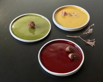 May include: Three small ceramic dishes with white rims. The dishes are filled with a glossy glaze in shades of green, yellow, and red. The green dish has a small brown acorn in the center. The red dish has two small brown acorns. The yellow dish has a small gold ring and a larger gold ring.