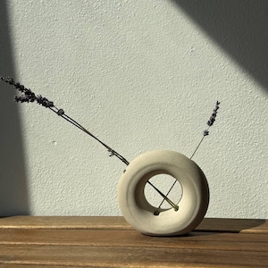 Könnte beinhalten: Eine kreisförmige, beige Keramikvase mit hohler Mitte und matter, texturierter Oberfläche. Zwei getrocknete Lavendelstiele stecken in der oberen Öffnung, wobei ein Stiel diagonal durch den zentralen Hohlraum des Gefäßes verläuft.