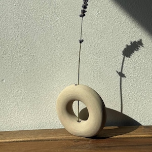 Könnte beinhalten: Eine kreisförmige, beige Keramikvase mit glatter, matter Oberfläche und einer zentralen Öffnung. Ein einzelner, schlanker Stängel aus getrocknetem violettem Lavendel steckt in der kleinen Öffnung oben an der Vase und ragt vertikal empor.