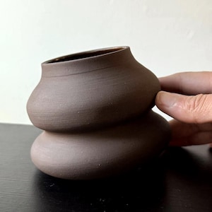 May include: A brown ceramic vase with a unique, stacked design. The vase has a wide, open top and a textured surface. The hand-thrown pottery is a dark, earthy brown color, perfect for displaying flowers or as a decorative piece.