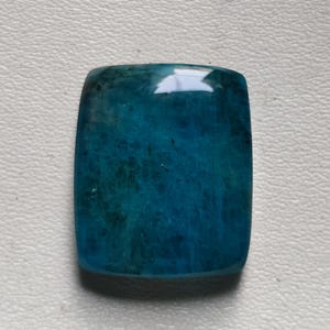 May include: A polished, rectangular gemstone with rounded corners. The stone is a deep teal colour with internal veining, and a glossy surface. The gemstone is set against a white background.