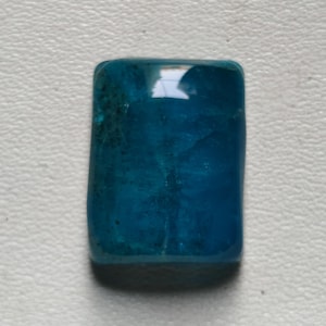 May include: A rectangular, polished gemstone in a deep teal hue. The stone has a smooth, glossy surface with subtle tonal variations, reflecting light. This gemstone is likely used in jewellery or as a decorative element.