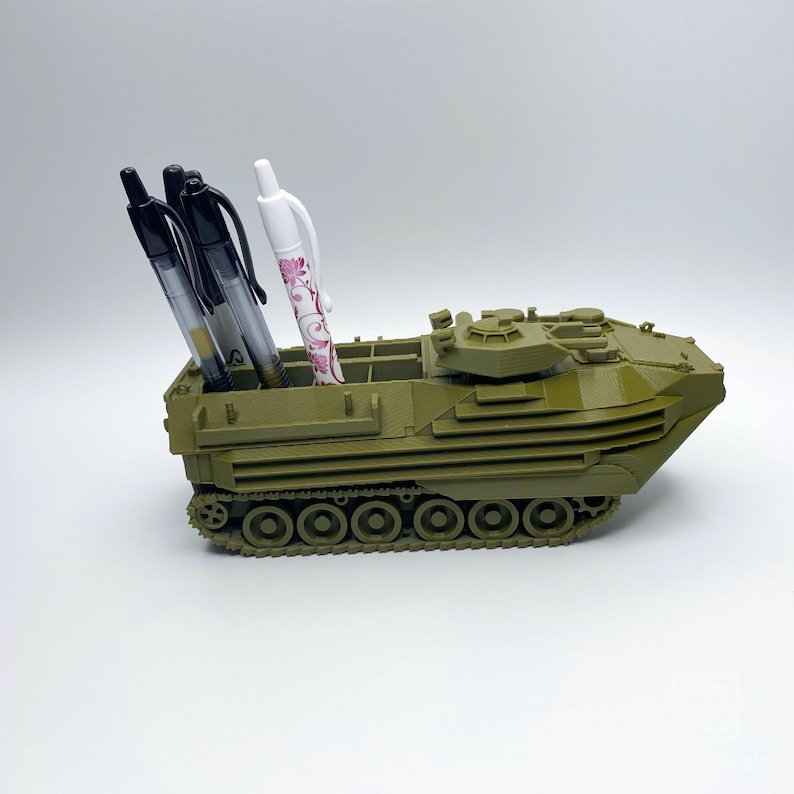 May include: A green toy military vehicle with a pen holder. The vehicle has a turret and tracks. There are three pens in the holder, one white and two black.