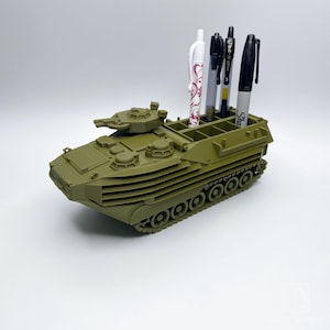 May include: A green plastic model of an amphibious assault vehicle with a pen holder on top. The pen holder has four slots for pens and pencils. The model has a detailed design with a turret and tracks.