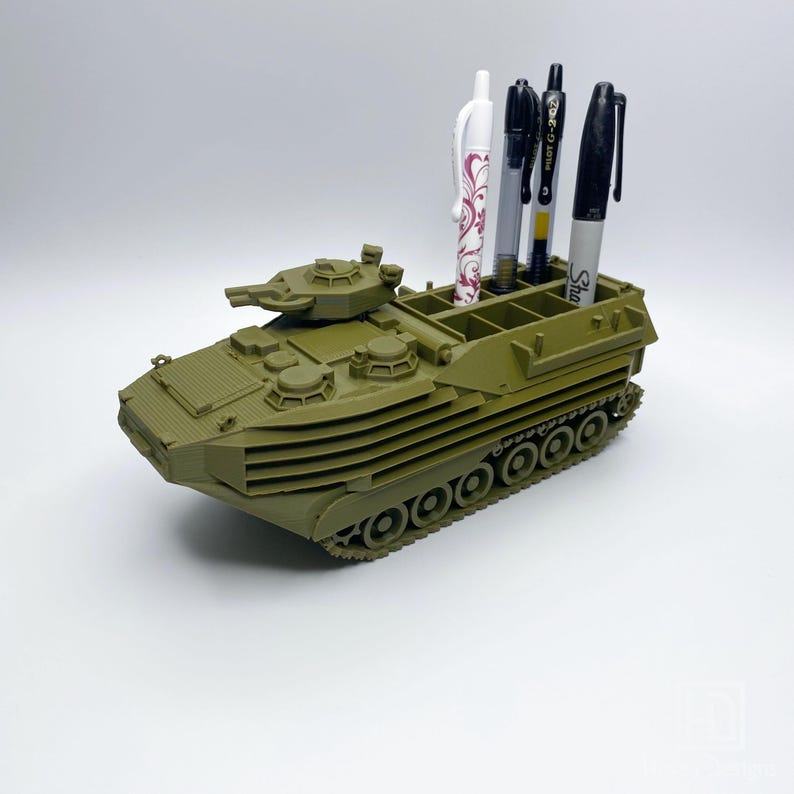May include: A green plastic model of an amphibious assault vehicle with a pen holder on top. The pen holder has four slots for pens and pencils. The model has a detailed design with a turret and tracks.