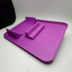 Puede incluir: Una bandeja rectangular de color morado que presenta una estructura de rampa elevada y un bloque elevado separado en la superficie. La bandeja tiene el texto TECH TRAY en relieve en el lado derecho junto a un icono gráfico de una campana con un ojo.
