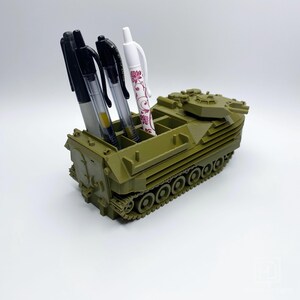 May include: A green toy military vehicle with a pen holder. The vehicle has a turret on top and a track system. There are four pens in the holder, including one with a floral design.