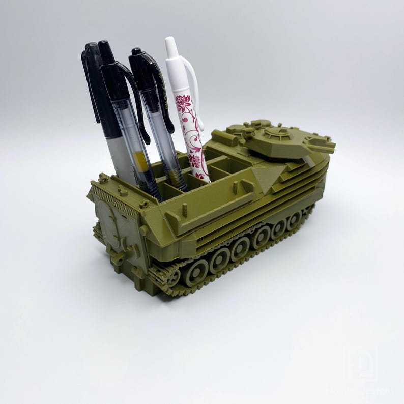 May include: A green toy military vehicle with a pen holder. The vehicle has a turret on top and a track system. There are four pens in the holder, including one with a floral design.
