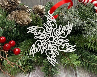 May include: A white snowflake-shaped ornament featuring the word Melody repeated in a script font, suspended by a red ribbon. The ornament is displayed against a background of green pine branches, gold-toned pinecones, and red berries.