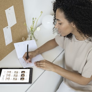 Puede incluir: Una persona escribe en una agenda en un escritorio blanco con un bol&iacute;grafo negro. Una tableta que muestra im&aacute;genes est&aacute; en el escritorio. Un corcho con bocetos y un jarr&oacute;n con flores est&aacute;n en el fondo. La persona lleva una camiseta beige.